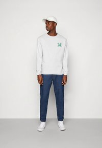 Sweatshirt cinzento de mangas compridas com um logótipo verde "M", combinado com jeans azul escuro e ténis brancos, completa o look casual.