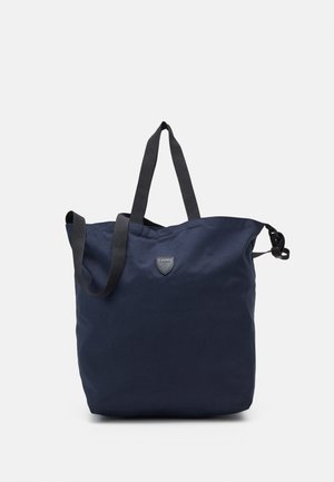 Sac cabas bleu marine avec poignées noires, petite étiquette logo à l'avant et détail boucle latérale sur fond blanc.