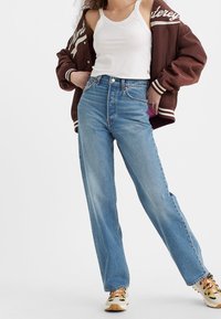 Jean taille haute bleu clair avec une coupe droite, en denim. Associé à un débardeur blanc et une veste de varsity marron.