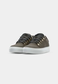 Barna bőr sneaker arany részletekkel, vastag szürke fűzőkkel, fehér gumitalppal és sima, texturált felülettel.