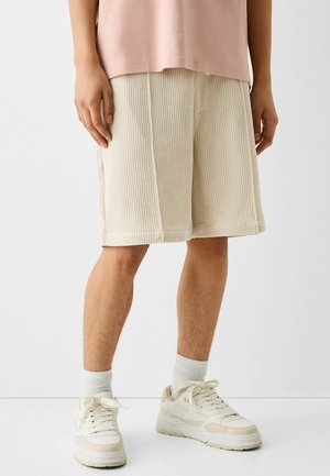 Osoba ubrana w kremowe prążkowane szorty do kolan, różową bluzkę, białe skarpetki i kremowe sneakersy, stojąca na tle jednolitego białego tła.