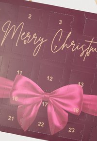 Μωβ ημερολόγιο Advent με χρυσά νούμερα, ροζ φιόγκο κατά μήκος του κέντρου και τη φράση «Καλά Χριστούγεννα» γραμμένη με χρυσά γράμματα καλλιγραφίας.