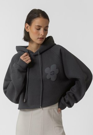 Sweat à capuche gris court en tissu doux, orné d'un grand patch floral sur le côté gauche et de cordons de serrage avec des embouts en métal.