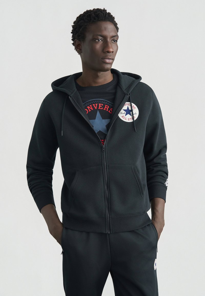 Sort lynlåshættetrøje lavet af bomuldsblanding med frontlomme, der har et rundt grafisk mærke på brystet og tilspidsede ærmekanter.