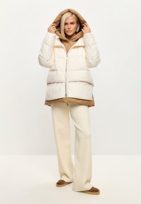 Puff-jakke i creme med en kontrasterende brun kant og hætte. Har en fuld lynlås og to frontlommer. Bæres over en beige hoodie og bukser.