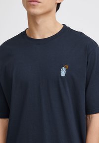 Navy blue cotton t-shirt featuring a small embroidered design of a beverage with an orange slice on the left chest area. Short sleeves.