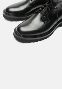 Scarpe in pelle nera con finitura lucida, punta tonda e design con lacci. Presentano una suola in gomma nera testurizzata e dettagli cuciti.