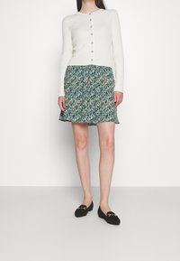 Cardigan long à manches longues côtelé blanc avec des boutons, associé à une jupe à motifs floraux teal avec un ourlet festonné, et des mocassins noirs avec des accents dorés.