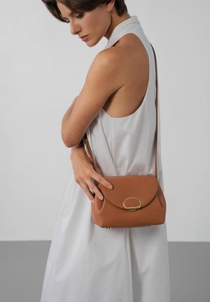 Femme en robe blanche sans manches tenant un sac à bandoulière en cuir marron avec une boucle dorée, sur un fond gris et blanc.
