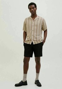 Chemise à manches courtes beige rayée avec des boutons, portée avec un short noir. Chaussures noires avec des chaussettes. Design simple et décontracté.
