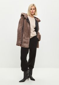 Chaqueta con capucha de piel sintética marrón y cremallera negra, llevada sobre un suéter beige y pantalones jogger negros. Botas negras con tacón completan el conjunto.