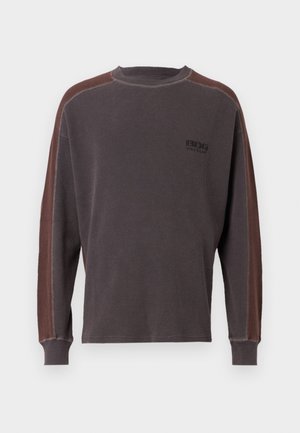 Tumšs pelēks garām piedurknēm krekls ar kontrastējošām brūnām sānu joslām. Tekstūrēta auduma, riboti piedurkņu gali un neliels logo uz krūtīm.