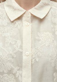 Col et devant de blouse blanche transparente avec une délicate broderie florale et de petits boutons ronds.