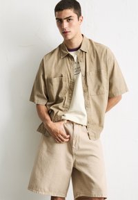 Beige Kurzarmhemd mit Knopfleiste und zwei Brusttaschen, kombiniert mit beigen Shorts. Glattes Material, lockere Passform, minimalistisches Design.