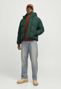 Grøn puffjakke med hætte, kviltet design, båret over en brun sweater, sammen med lyseblå jeans og beige støvler. Brun cap på hovedet.