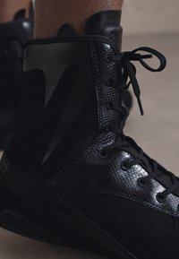 Scarpa da ginnastica alta nera con tomaia in mesh testurizzato, dettagli lisci e lacci piatti, progettata per le prestazioni e il supporto.