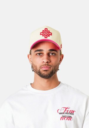 Man met verzorgde baard die een crèmekleurige pet met rood dessin draagt en een wit T-shirt met de tekst "Fear none" in rood en zwart.