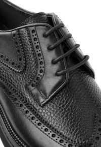 Schwarzer Leder-Brogue-Schuh mit Wingtipp-Perforationen, strukturierten Mustern und geschlossener Schnürung im oberen Bereich.