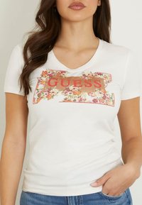 Biała bawełniana koszulka z dekoltem w serek i krótkimi rękawami z kwiatowym wzorem oraz napisem "GUESS" w odważnym czerwonym tekście z przodu.
