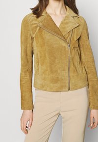 Tan suede motorcycle jacket with an asymmetrical zipper, long sleeves, and snap collar. Features a fitted cut and two side pockets.