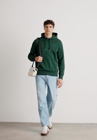Felpa verde con cordini e logo, abbinata a jeans azzurri chiari e sneakers bianche; porta una piccola borsa a tracolla color crema.