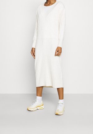Lange, geribbelde gebreide jurk in off-white, met een V-hals en lange mouwen. Gecombineerd met lichte sneakers, wat zorgt voor een casual uitstraling.