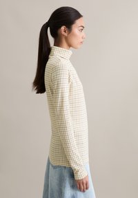 Dopasowany top z długim rękawem i golfem, wykonany z miękkiej kremowej tkaniny z drobnym brązowym wzorem w kratkę. Lekka faktura, prosty dół.