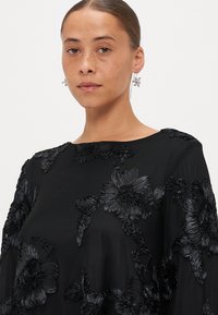 Melna ziedu izšūta blūze, kurā ir trīsdimensiju melni ziedi un lapas, ar apaļu kaklu un mīkstu, plūstošu audumu.