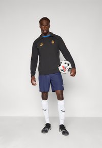 Sort langærmet sportstop med orange accenter, marineblå shorts, hvide sokker med sort logo, holder en sort-hvid fodbold.