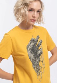 Gele katoenen t-shirt met een zwarte afbeelding van een hand met draad en naalden, met een gladde textuur en een klassieke ronde halslijn.