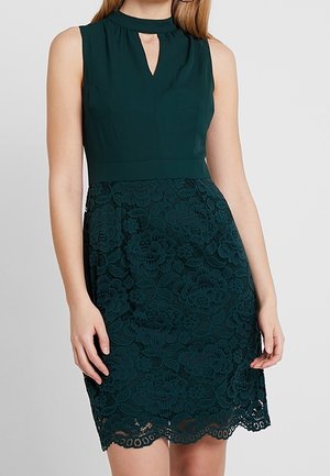 Femme portant une robe verte foncée sans manches avec un décolleté en trou de serrure et une jupe en dentelle florale, debout sur un fond clair uni.