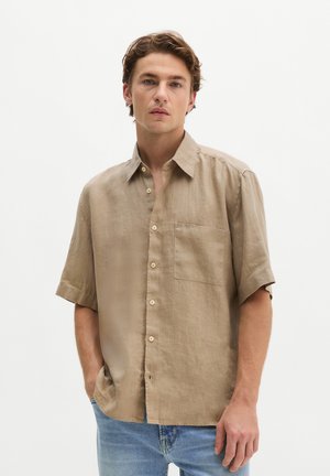 Junger Mann mit welligem braunem Haar, der ein beiges kurzärmeliges Hemd und blaue Jeans trägt, steht mit einer Hand in der Tasche.