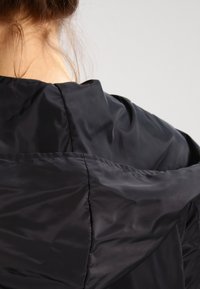 Černá, lehká bunda s hladkým, lesklým povrchem. Obsahuje kapuci a viditelné švy na zadní straně a ramenou.