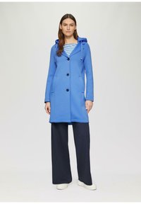 Cappotto blu con cappuccio, dalla texture liscia, maniche lunghe, bottoni frontali e due tasche laterali, indossato sopra una maglietta a righe e pantaloni ampi scuri.