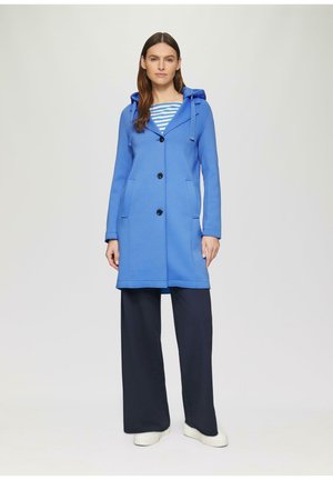 Cappotto blu con cappuccio, dalla texture liscia, maniche lunghe, bottoni frontali e due tasche laterali, indossato sopra una maglietta a righe e pantaloni ampi scuri.