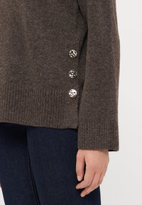 Brun ullgenser med ribbet kant, med tre dekorative knapper på ermet. Glatt tekstur, avslappet passform. Blå jeans synlige under.