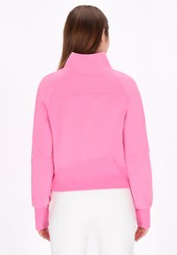 Fleecepullover i stark rosa med hög krage, raglanärmar och ribbade ärmslut. Slät textur med sömdetaljer på baksidan.