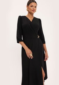 Czarna sukienka kopertowa, z rękawami o długości trzech czwartych, z wcięciem w talii z metalicznym akcentem oraz rozcięciem na udzie dla dodatkowego detalu.