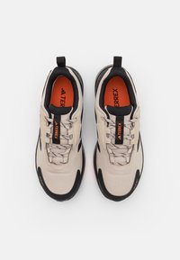Beżowe buty sportowe z czarnymi akcentami, teksturowana tkanina, szare sznurowadła, pomarańczowa wkładka oraz pętelki z tyłu i z przodu.