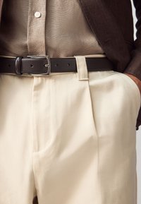 Béžové nohavice na mieru s prednými záhybmi, doplnené tmavohnedým textúrovaným opaskom so striebornou sponou a svetlohnedou košeľou pod nimi.