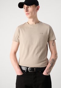 Μπεζ βαμβακερό μπλουζάκι με στρογγυλή λαιμόκοψη, κοντά μανίκια και μια μικρή λογότυπη λεπτομέρεια στο στήθος, σε συνδυασμό με μαύρα τζην και μαύρο καπέλο.