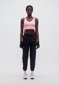 Γυναίκα όρθια, φοράει ροζ αθλητικό σουτιέν, μαύρη φόρμα, μαύρα γάντια χωρίς δάχτυλα, λευκές κάλτσες και ροζ αθλητικά παπούτσια σε λευκό φόντο.