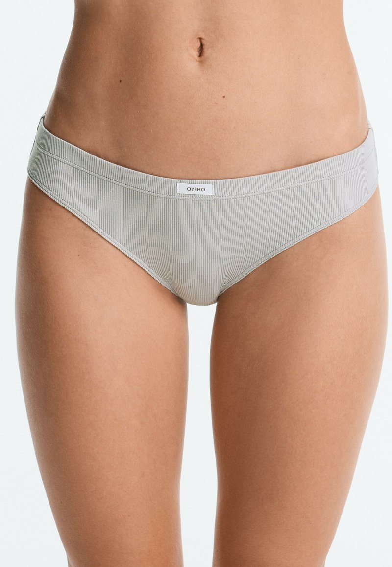 Femme portant un sous-vêtement style bikini côtelé gris clair avec une petite étiquette frontale sur un fond uni.