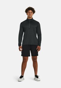 Schwarzes langärmeliges Shirt mit Viertelreißverschluss, kombiniert mit schwarzen Shorts, getragen mit weißen Sportschuhen mit schwarzen Akzenten, vor einem weißen Hintergrund.