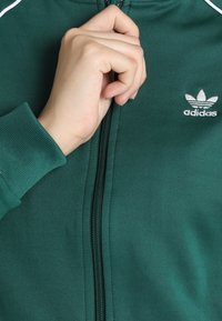 Zelená bunda na zip z hladké tkaniny s stříbrným logem Adidas a vroubkovanými manžetami. Zip vede vertikálně, má přiléhavý límec.