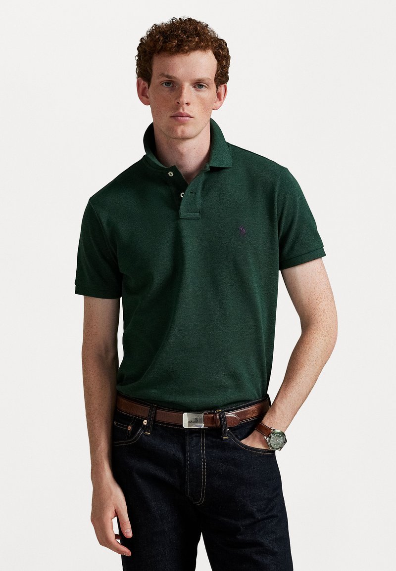 Groene polo met een drieknopenkraag, korte mouwen en een klein paars logo op de borst. Draag het met donkere jeans en een bruine leren riem.
