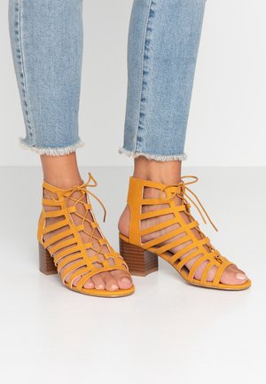 Geel leren vetersandalen met hak, met een gestructureerd ontwerp, open teen en blokhak, voorzien van een houten textuur en enkelbandjes.