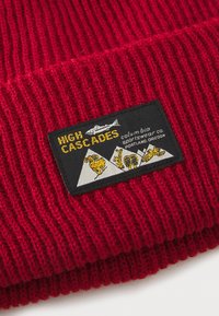 Červená pletená čepice s žebrovanou strukturou. Obsahuje obdélníkovou černou nášivku s grafikou hor v několika barvách a textem "HIGH CASCADES."