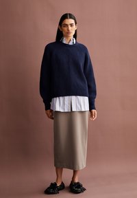 Marineblauwe gebreide trui met verlaagde schouders, gedragen over een witte gestreepte blouse, gecombineerd met een beige midi-rok en zwarte schoenen met franjes.
