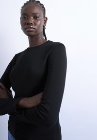 Αυτοπεποίθηση νέα γυναίκα με πλεκτά μαλλιά, που φοράει κρίκους στα αυτιά και μαύρη μακρυμάνικη μπλούζα, στέκεται με σταυρωμένα τα χέρια μπροστά σε λευκό φόντο.